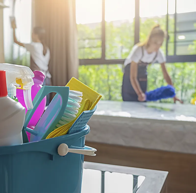 A blue bucket filled with cleaning supplies in the foreground, with CBM Smart Services professionals performing a bright Airbnb turnover cleaning in Massachusetts in the background.