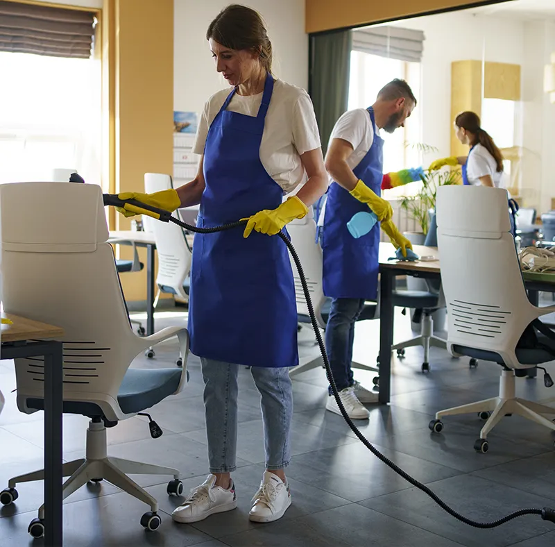 CBM Smart Services professional cleaners performing commercial office cleaning in Greater Boston, Massachusetts, vacuuming an office chair and wiping desks.
